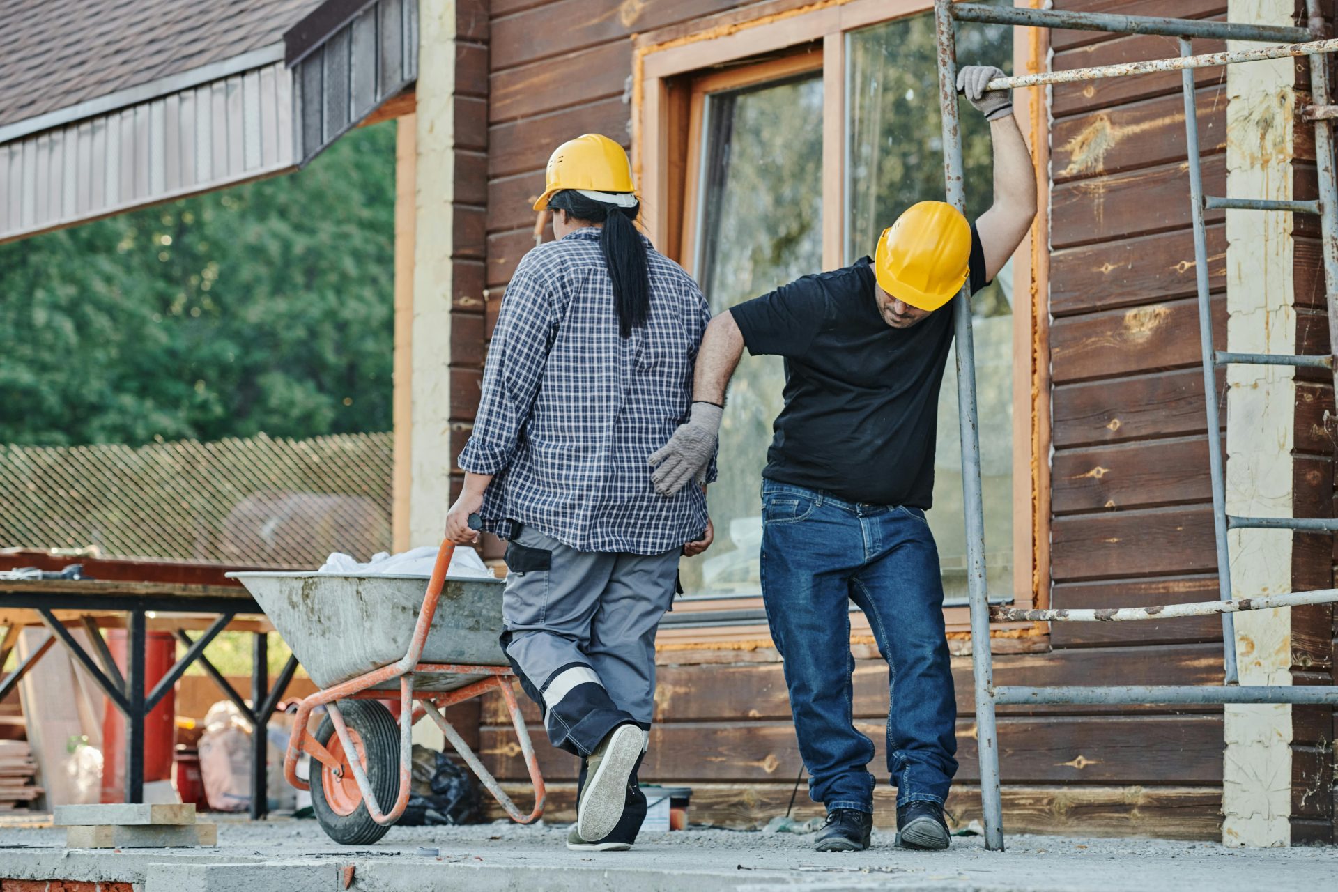 Two construction workers wearing hard hats work at an outdoor site, one with a wheelbarrow.
