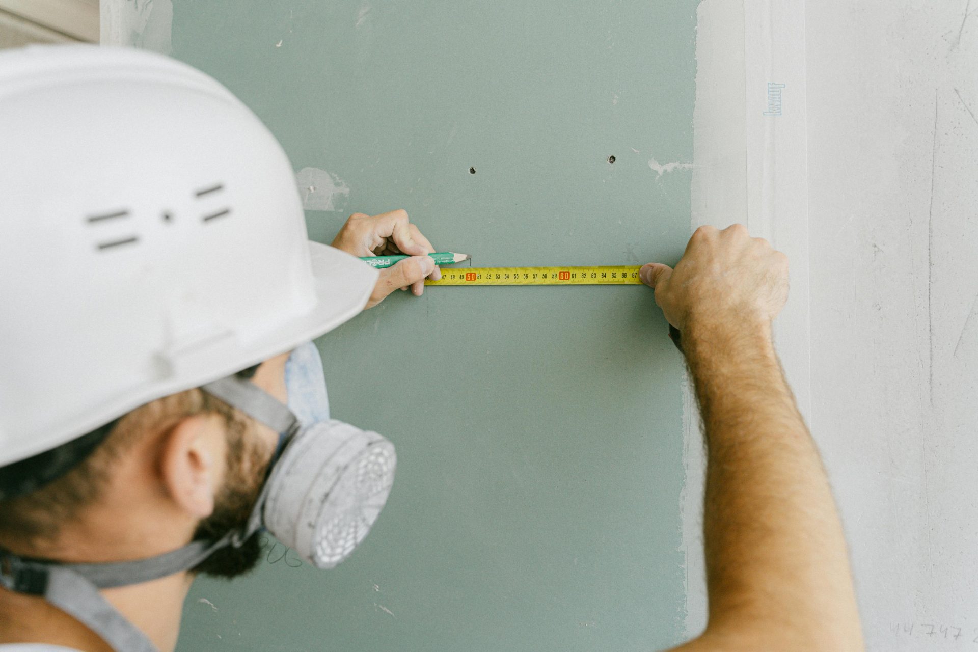 Ein Bauarbeiter, der präzise Gipskartonplatten ausmisst und dabei zu seiner Sicherheit einen Helm und eine Maske trägt.