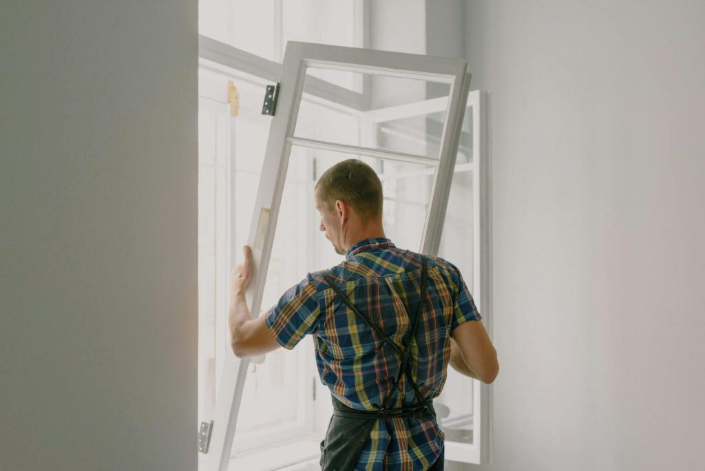 Rückansicht eines anonymen männlichen Monteurs in kariertem Hemd und Schürze, der zu Hause ein Fenster mit Scharnieren an einer weißen Wand montiert.
